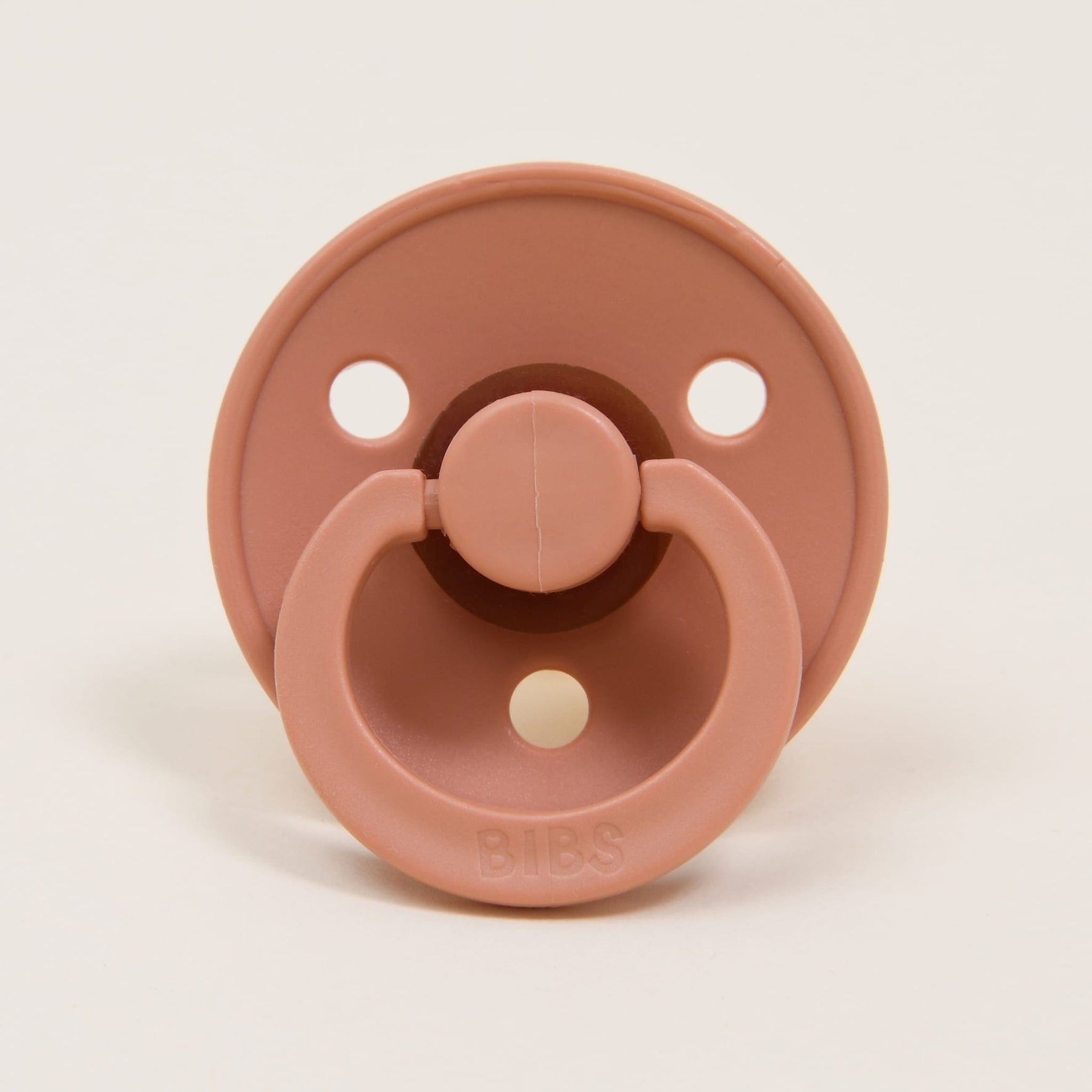 A round pacifier in a peach sunset color and crafted from natural rubber features two holes on the shield and a handle embossed with "BIBS." Designed in Denmark, this pacifier encourages babies to self-soothe. The backdrop is a plain, light color.