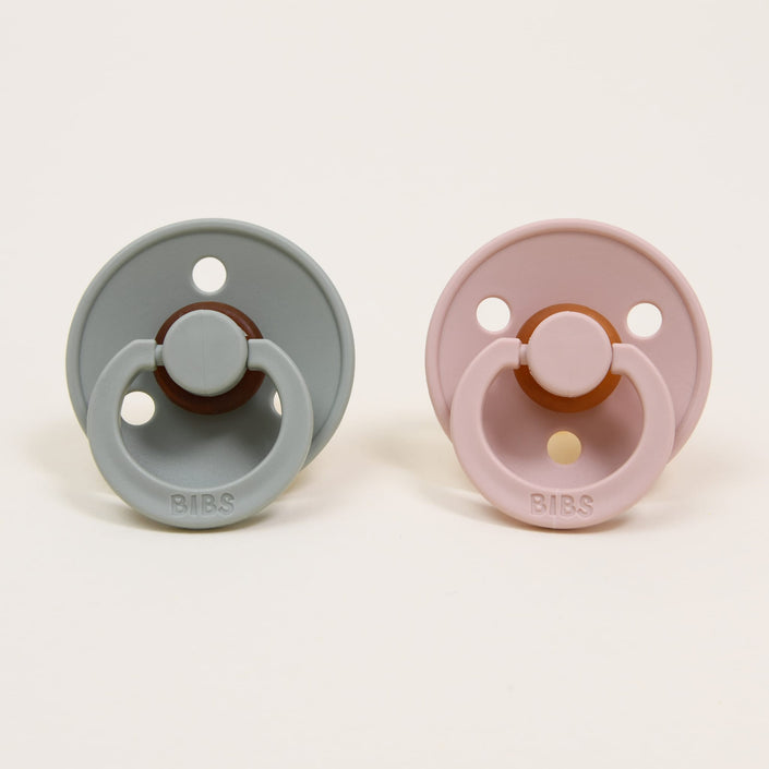 In the image, a set of two BIBS pacifiers crafted from natural rubber in Denmark is displayed against a plain background. On the left, the sage-colored pacifier complements the blush one on the right. Both feature round shields with two holes, encouraging babies to self-soothe, and proudly display the embossed "BIBS" brand name at their base.