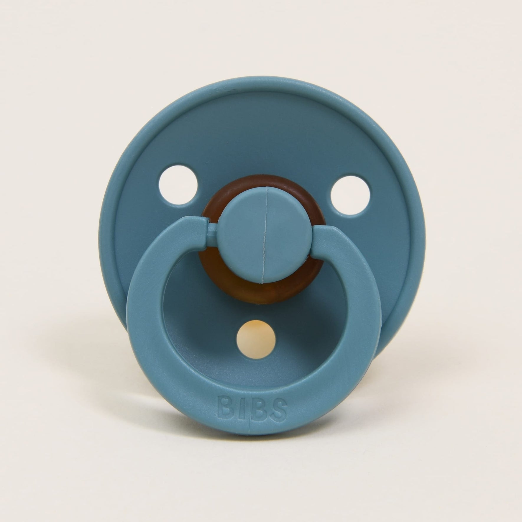 This Bibs Pacifier, in a classic and sophisticated Island Sea shade, is crafted from natural rubber. It boasts a rounded shield with two large holes and an embossed "BIBS" logo on the handle. Designed to aid babies in self-soothing, this pacifier stands upright against a neutral backdrop.