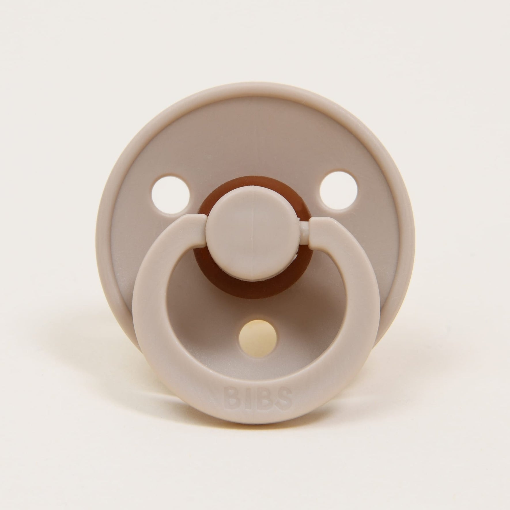 A timeless and elegant vanilla-colored round natural rubber baby pacifier, featuring two ventilation holes and a single ring handle, is centered on a plain white background. The word "BIBS" is embossed on the handle, offering an ideal tool for little ones to self-soothe.