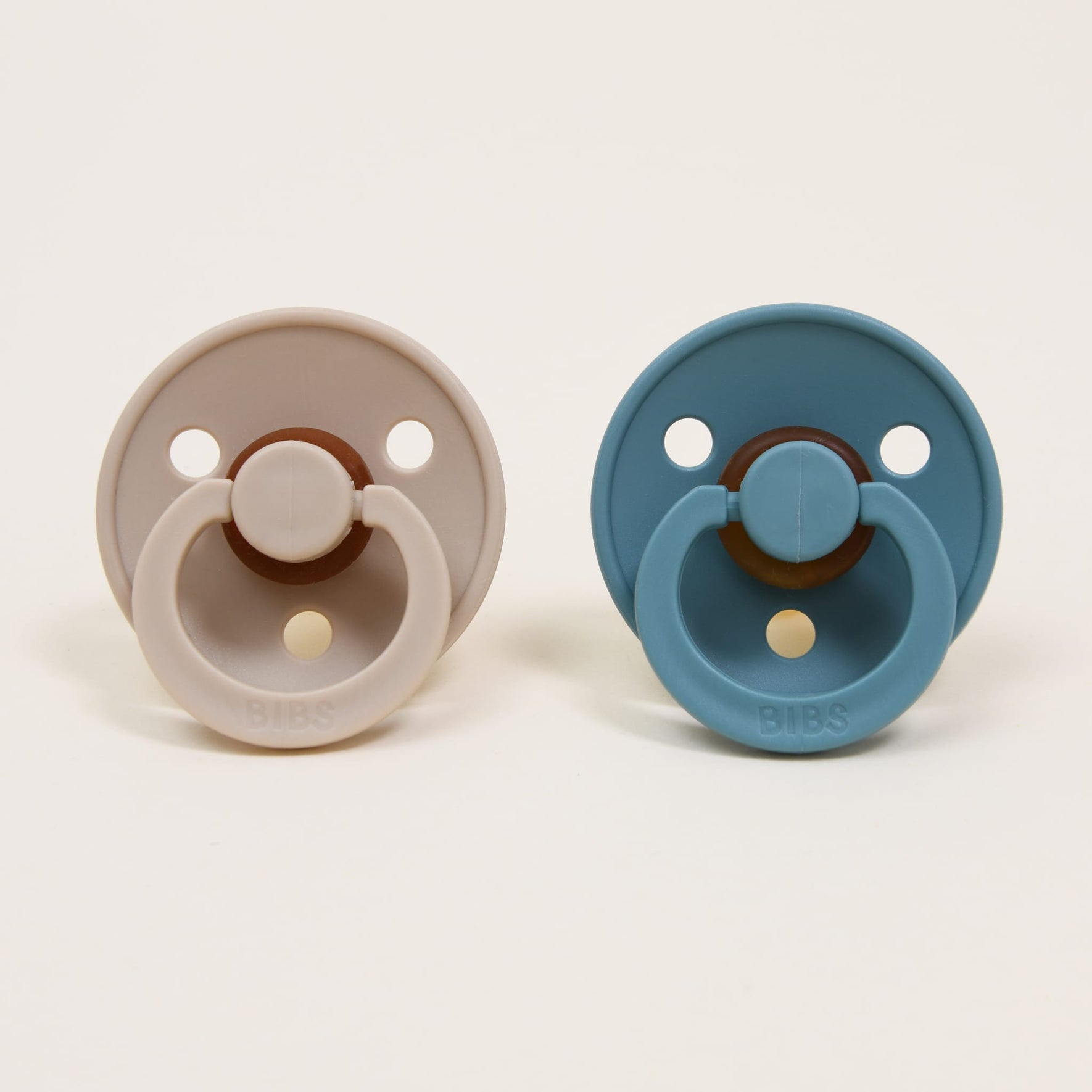 On a white background, two Bibs pacifiers are placed side by side. One is in vanilla and the other in island sea, both crafted from natural rubber. Each pacifier boasts a round shield with two ventilation holes and a circular handle embossed with "BIBS," offering a timeless and elegant touch that helps your little one self-soothe effortlessly.