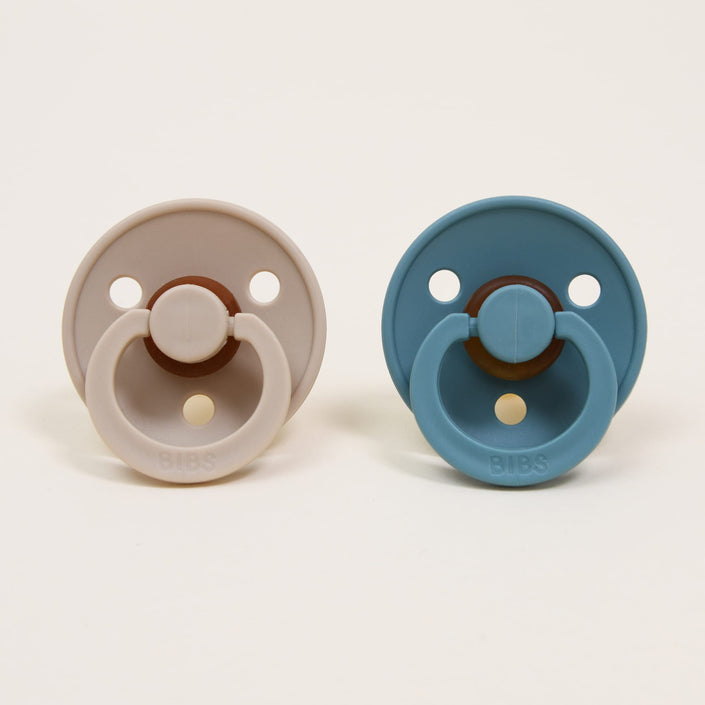 On a white background, two Bibs pacifiers are placed side by side. One is in vanilla and the other in island sea, both crafted from natural rubber. Each pacifier boasts a round shield with two ventilation holes and a circular handle embossed with "BIBS," offering a timeless and elegant touch that helps your little one self-soothe effortlessly.