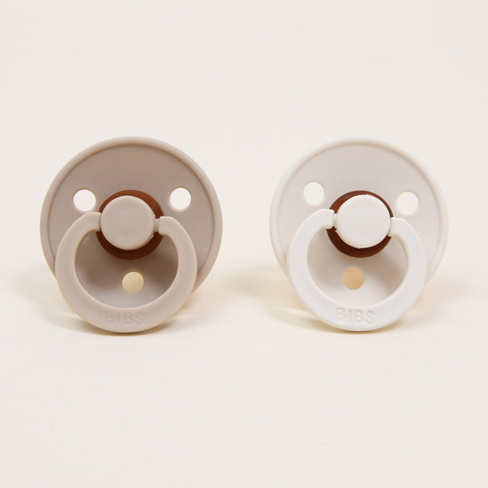 Displayed against a plain background are two round pacifiers from the Bibs Pacifier Set in Vanilla & Ivory. The pacifier on the left is light brown with a beige handle, while the one on the right is white with a matching handle. Both feature circular shields with two holes, showcasing Denmark's minimalist design to aid babies in self-soothing.