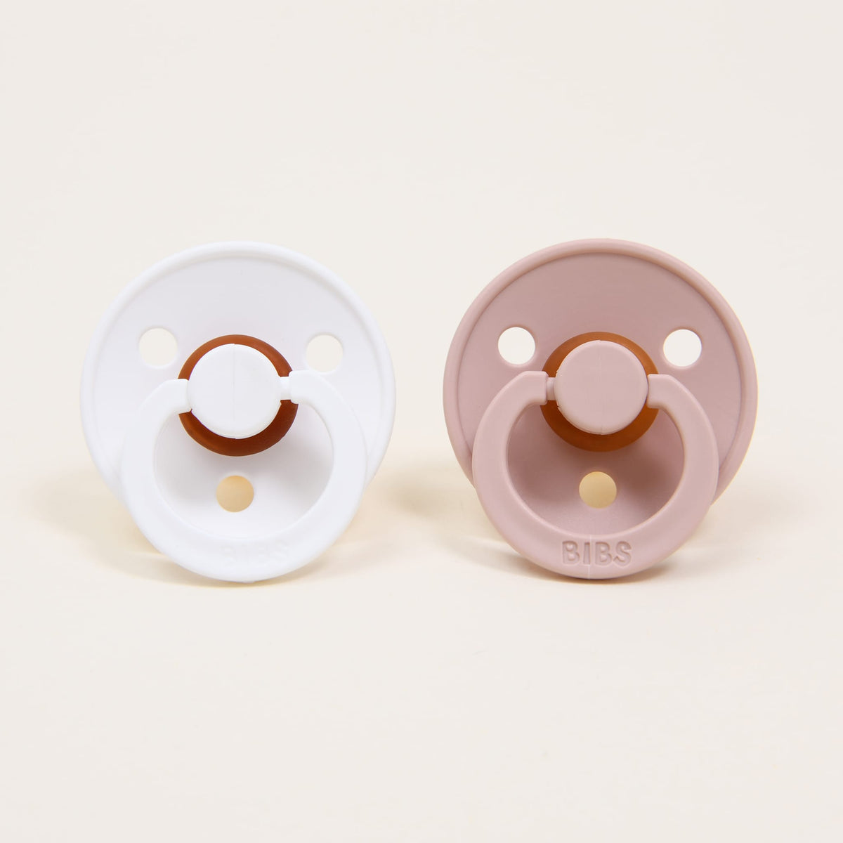 The Bibs Pacifier Set, featuring two pacifiers made from natural rubber to aid in baby self-soothing, is displayed against a plain background. The pacifier on the left is white, while the one on the right is blush, showcasing a circular design with two holes and a central button.