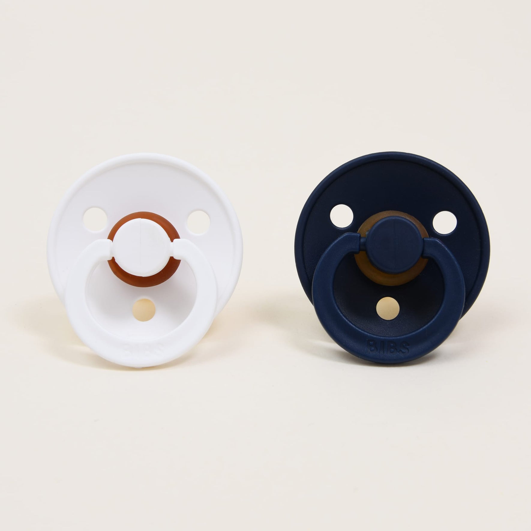 The Bibs Pacifier Set in White & Deep Space showcases two round pacifiers made from natural rubber against a neutral background. One pacifier features a white shell with a brown center, while the other displays navy blue and brown tones, creating a classic look. Both pacifiers include handles and are designed with several small holes to offer self-soothing comfort.