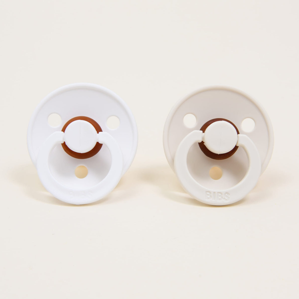 The Bibs Pacifier Set, made in Denmark from natural rubber, includes two pacifiers resting on a neutral background. Each pacifier is round and showcases calming white and brown tones. The left pacifier has a pristine white handle, while the right one presents a subtle ivory shade, perfect for self-soothing.