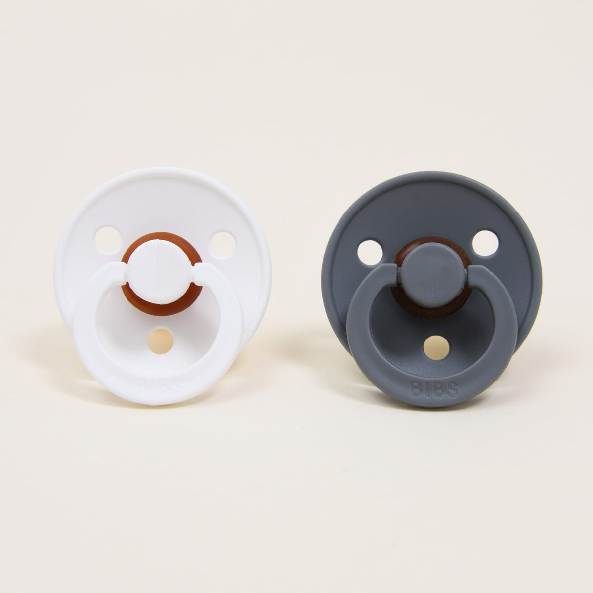 Displayed upright against a plain background are two round natural rubber pacifiers from the Bibs Pacifier Set in White & Smoke. Both pacifiers boast a classic design with circular guards, each featuring a central handle and ventilation holes.