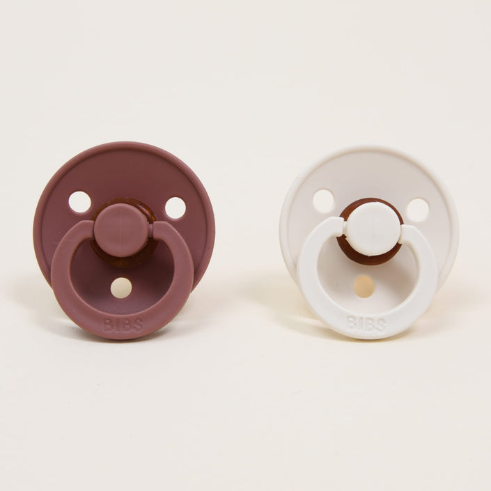 The Bibs Pacifier Set, featuring the Woodchuck and Ivory designs, showcases two pacifiers set against a neutral background: the dark red Woodchuck on the left and the timeless, elegant white Ivory on the right. Both pacifiers have a self-soothing design with a round shield and circular handle for comfort and are made from natural rubber.