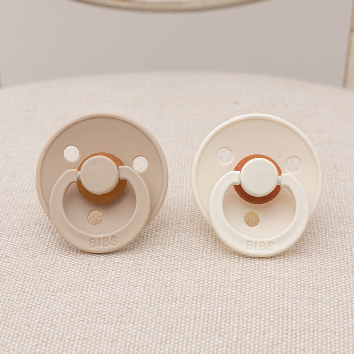 The Braden Pacifier Set in Vanilla and Ivory is shown on a textured surface. One pacifier is vanilla and the other is ivory, both featuring circular guards with two ventilation holes and branded as "BIBS." These natural rubber pacifiers have a classic design that assists infants in soothing themselves comfortably.