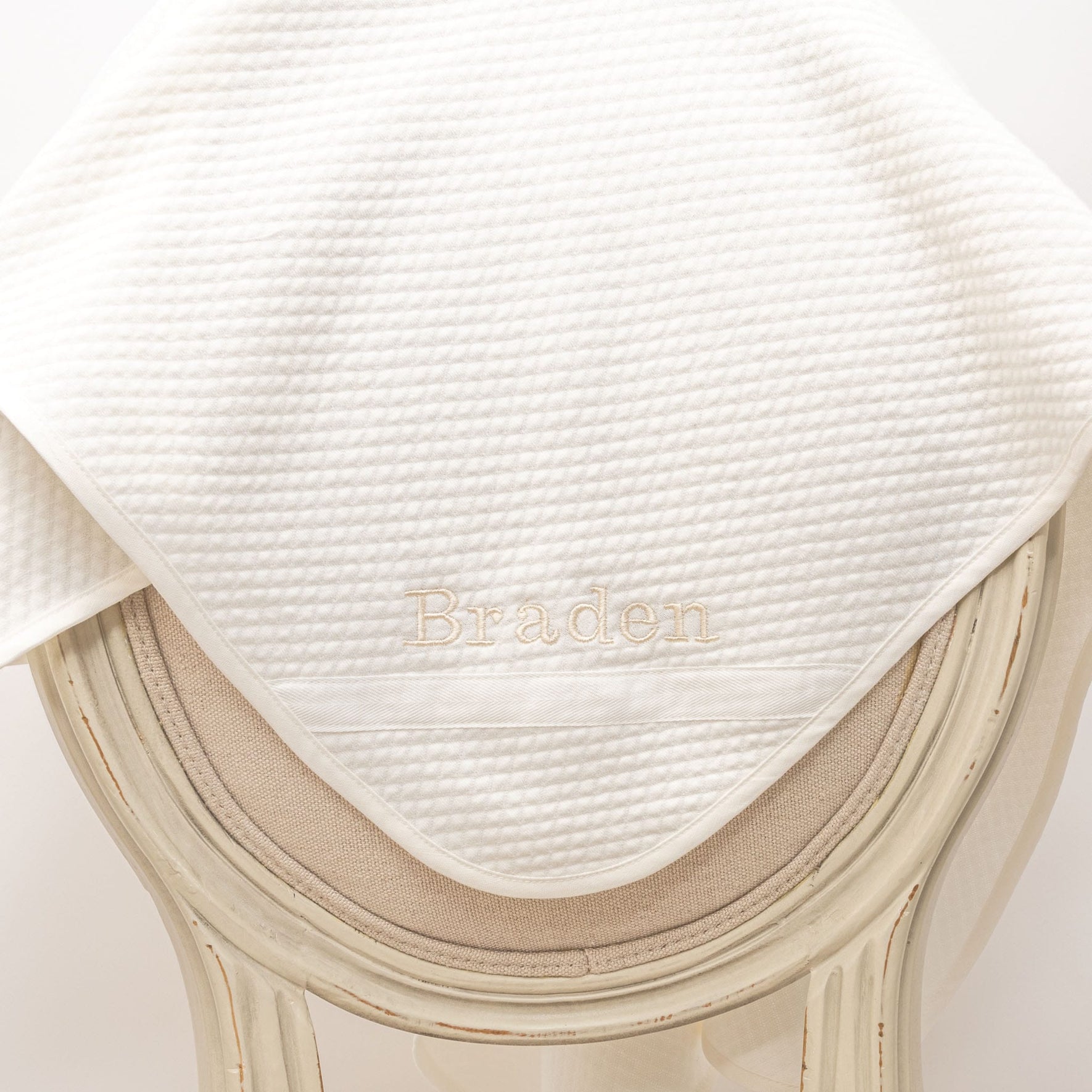 Flat lay photo of the Braden Personalized Blanket made from ivory textured cotton with an ivory grosgrain ribbon woven on the corner. The name "Braden" is embroidered on the corner of the blanket.