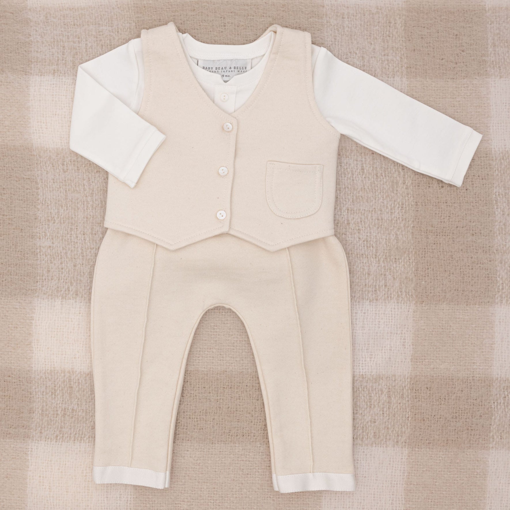 The Braden Vest Suit is a charming beige baby outfit perfect for any occasion. It features a long-sleeved white shirt paired with a beige vest adorned with buttons and a small pocket, along with matching pants. It is elegantly showcased on a soft, textured French Terry checkered background in beige and white.