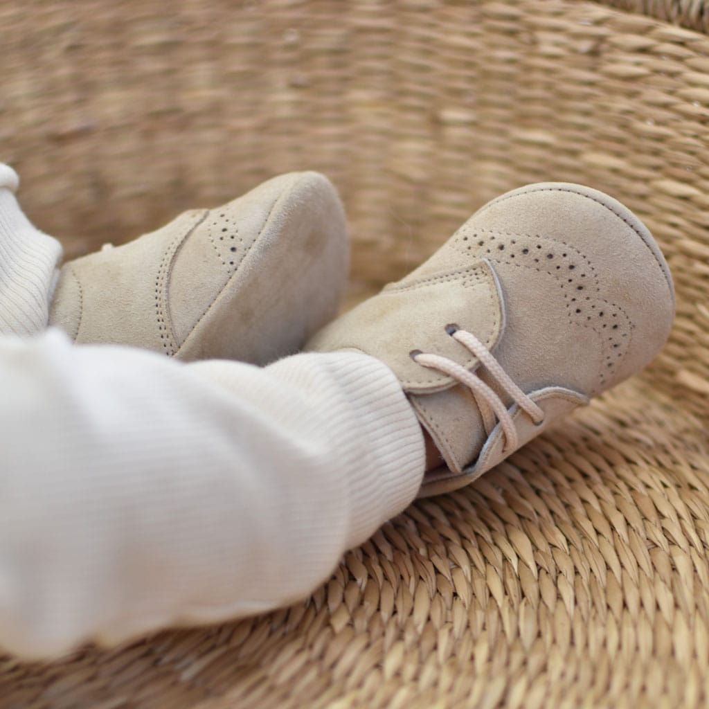 Close-up of a baby wearing Camel Suede Shoes from Baby Beau & Belle, resting on a woven basket. The baby is dressed in white knit pants. The handmade boys' shoes have decorative perforations on the sides and laces.