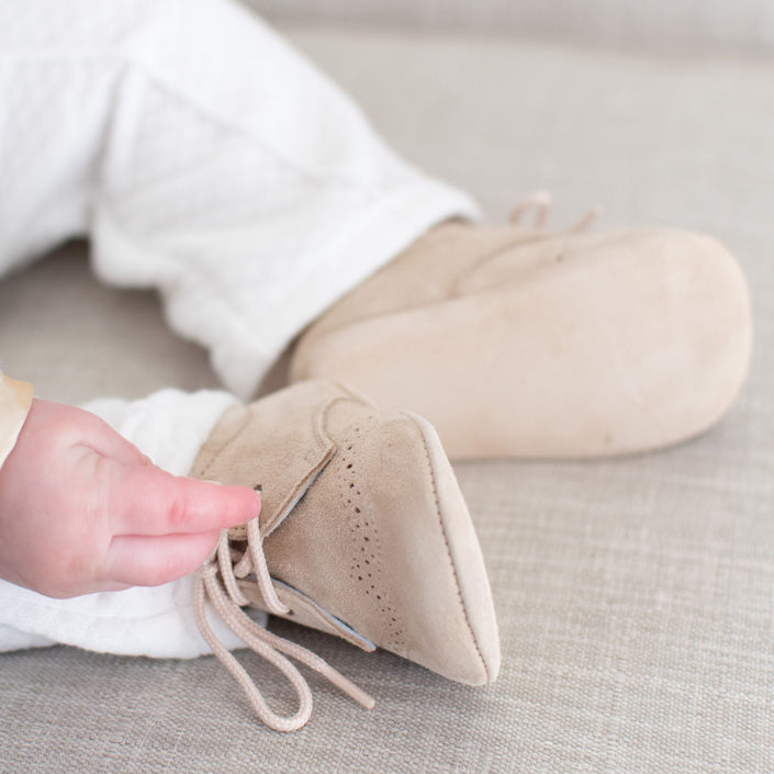 A close-up image displays a baby touching one of his light beige Camel Suede Shoes with laces. The baby, seated on a light-colored fabric surface, is dressed in white quilted pants.
