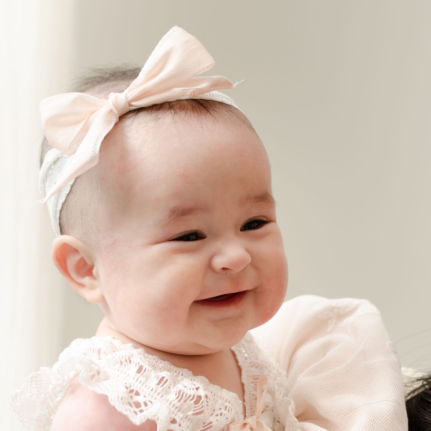 A baby is smiling while wearing a Charlotte Lace Headband. The baby is dressed in a white, floral lace-trimmed outfit and appears to be looking at someone off-camera. The background is a light, neutral color.
