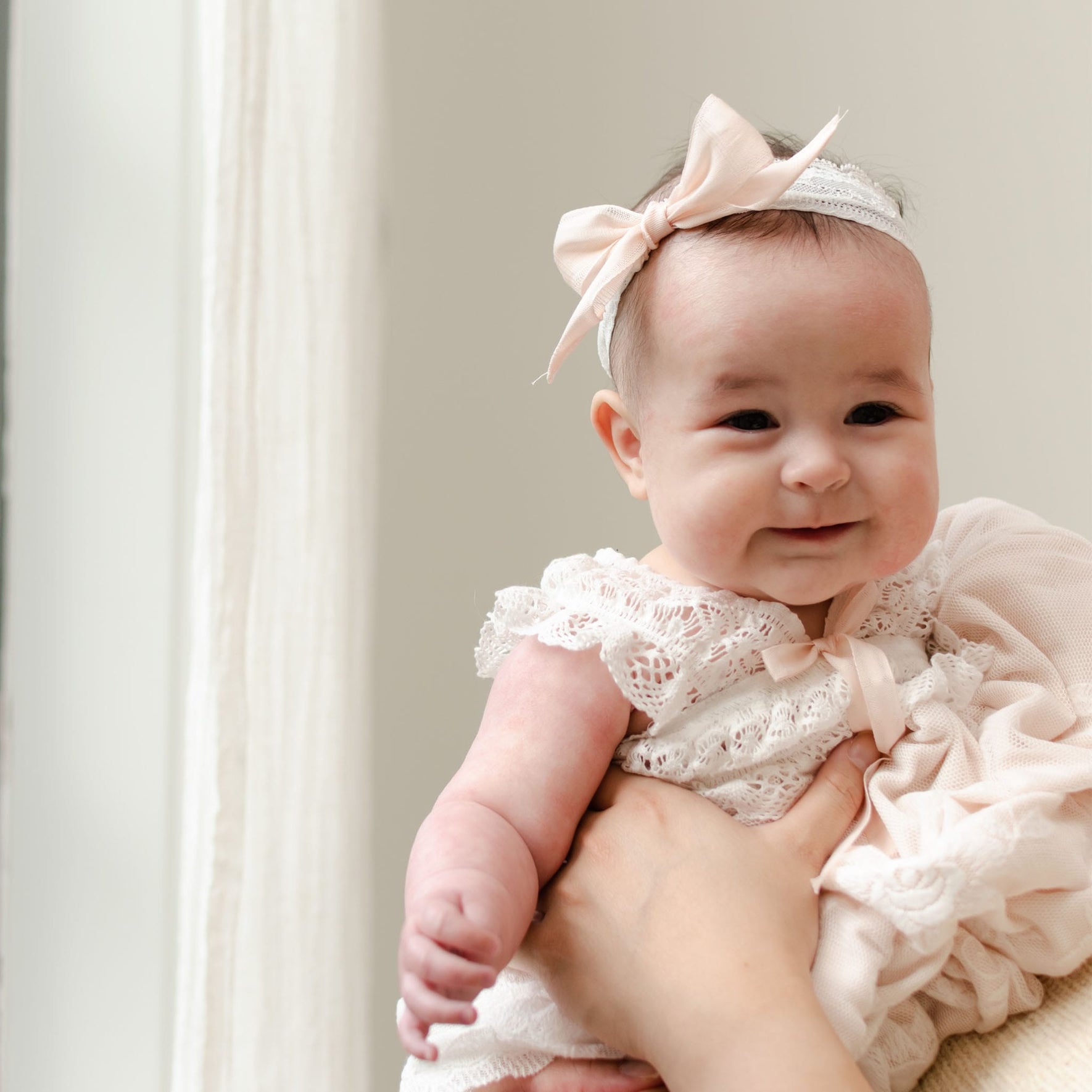 A baby wearing a Charlotte Lace Headband and a white floral lace outfit with a blush cotton ribbon is being held by an adult. The baby is smiling and looking at the camera, while the background shows a light-colored curtain.