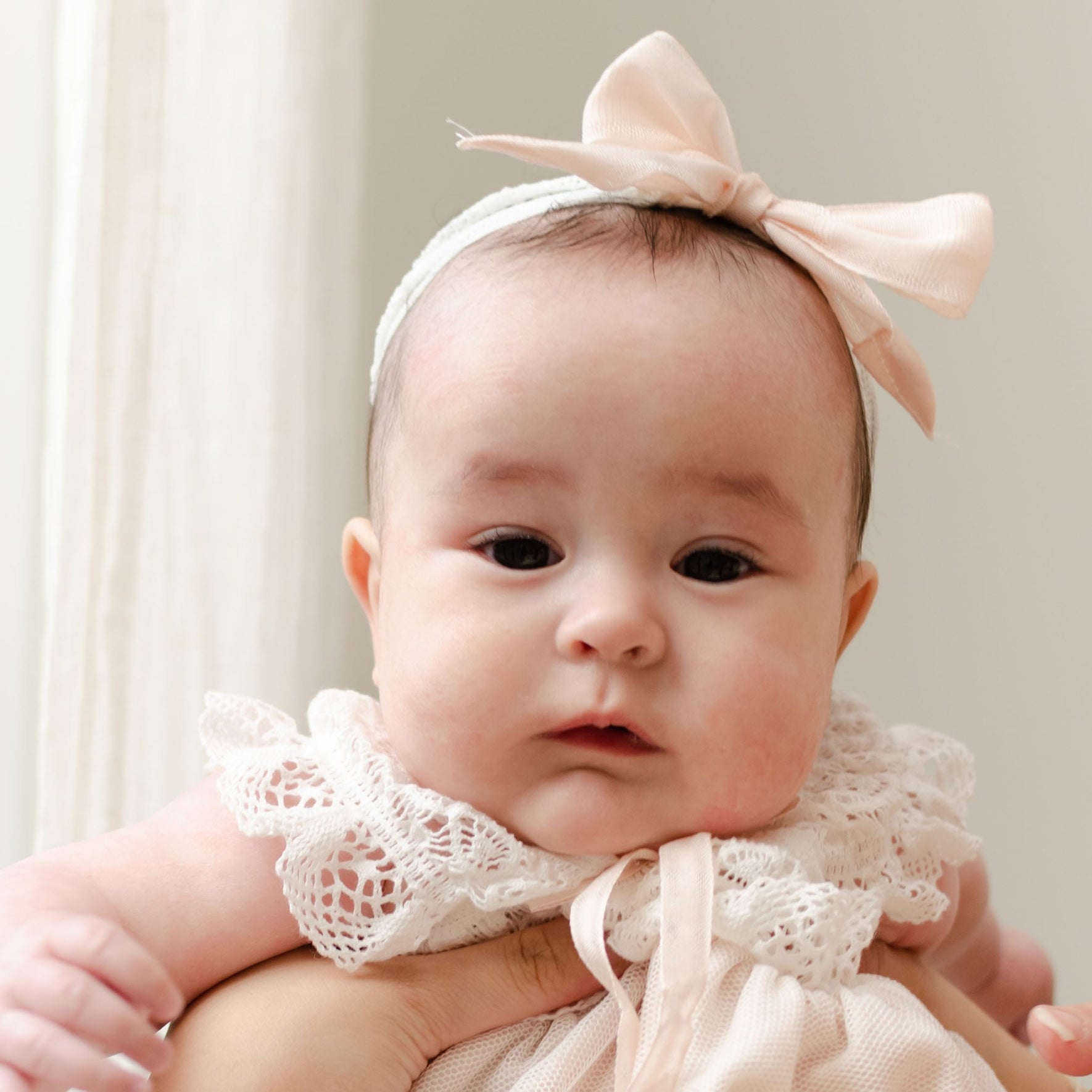 A baby dressed in a lacy white outfit and a large pale pink Charlotte Lace Headband on their head is being held up by an adult hand. The background is neutral and light, possibly a room with white walls. The baby has a calm expression and dark hair.