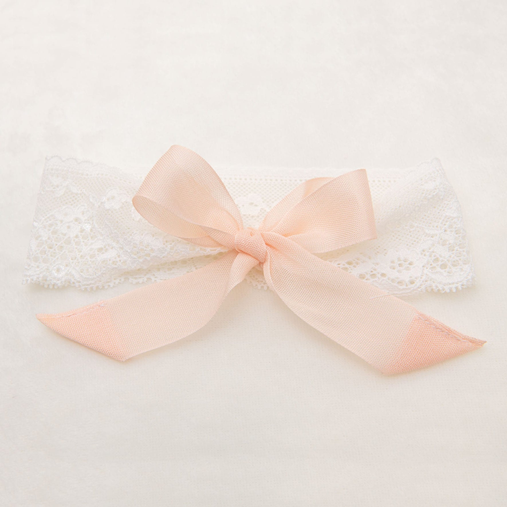 A Charlotte Lace Headband with a decorative pink bow is laid on a light, neutral background. Featuring an intricate floral lace pattern, the headband's blush cotton ribbon bow is neatly tied in the center, adding a charming touch.