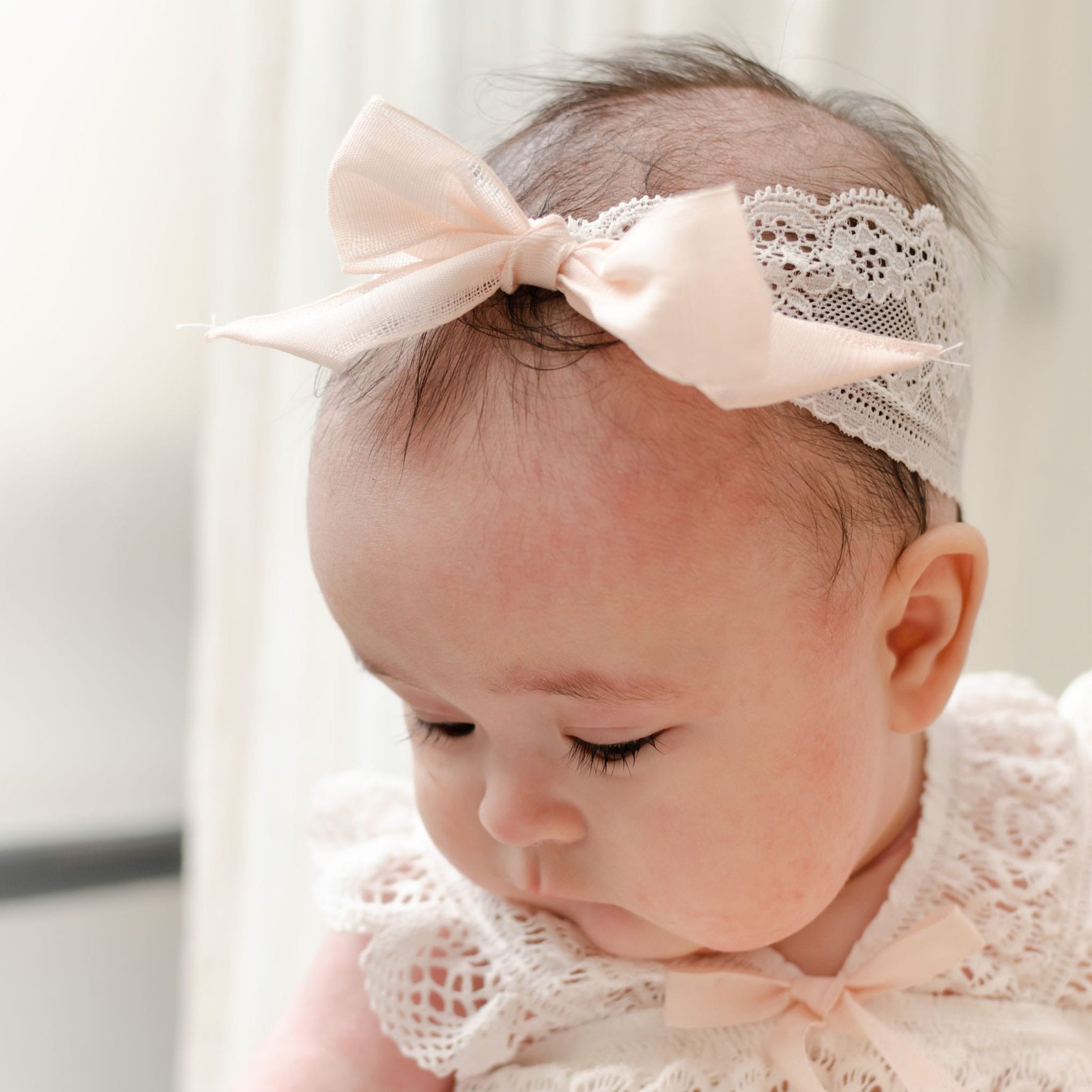 A close-up of a baby wearing a Charlotte Lace Headband with a blush cotton ribbon and a white lace dress. The baby is looking downward with closed eyes, and the background is softly blurred, suggesting an indoor setting with natural light.