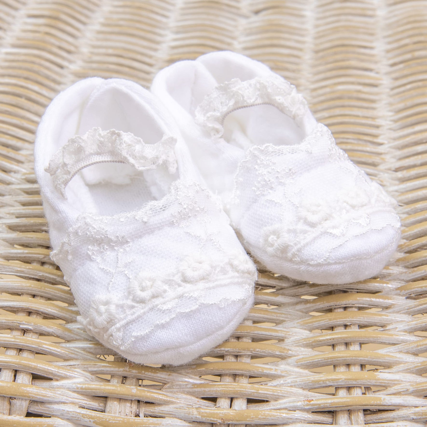 A pair of white baby shoes with lace details from the Eliza Accessory Bundle - Save 15%, perfectly complementing a baby dress accessory set, placed on a woven wicker surface.