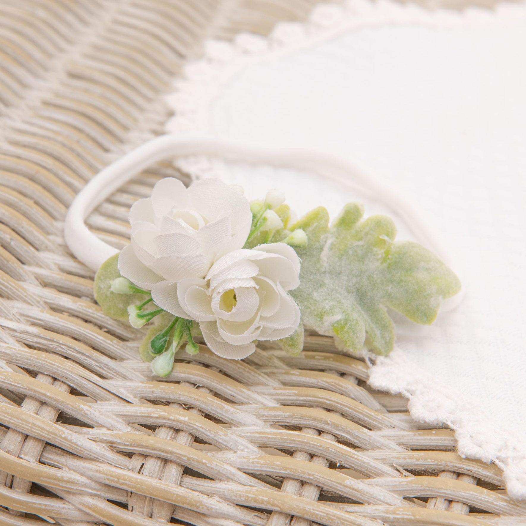 An Eliza Accessory Bundle, featuring a white fabric headband adorned with a petite artificial white rose and green leaves, rests on a woven rattan surface, while a personalized quilted blanket is partially visible in the background.