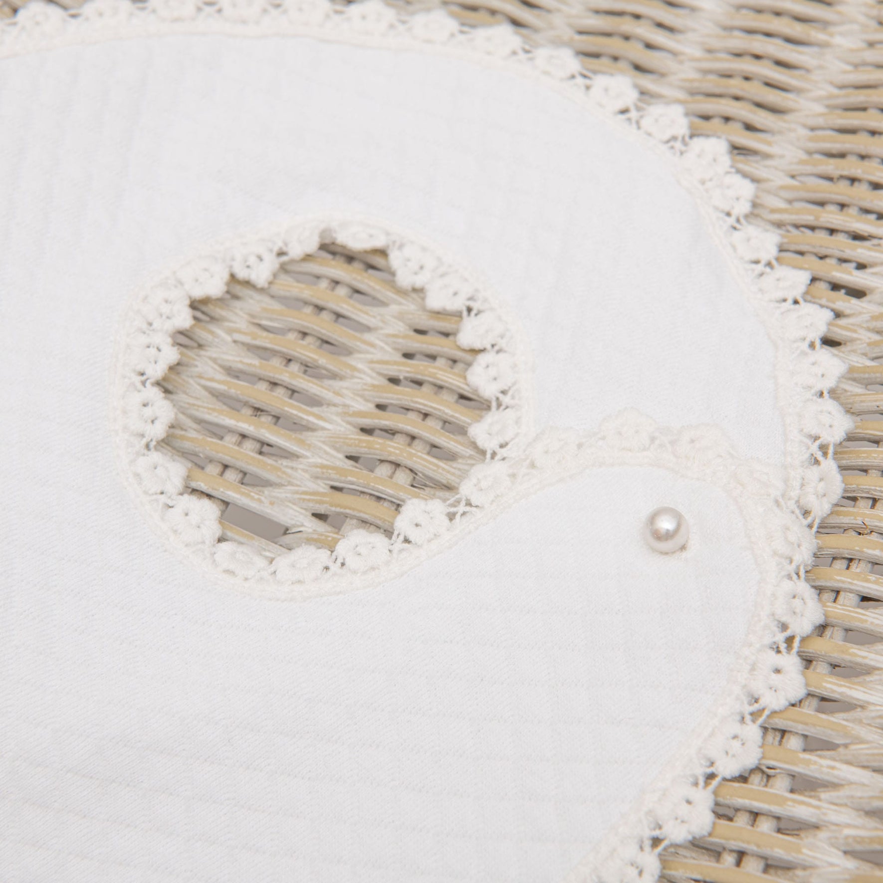 An Eliza Bib, a white, quilted fabric baby bib with crocheted floral lace trim, shaped like a bird, lays on a woven wicker surface. It has a small pearl-like button near the neckline.