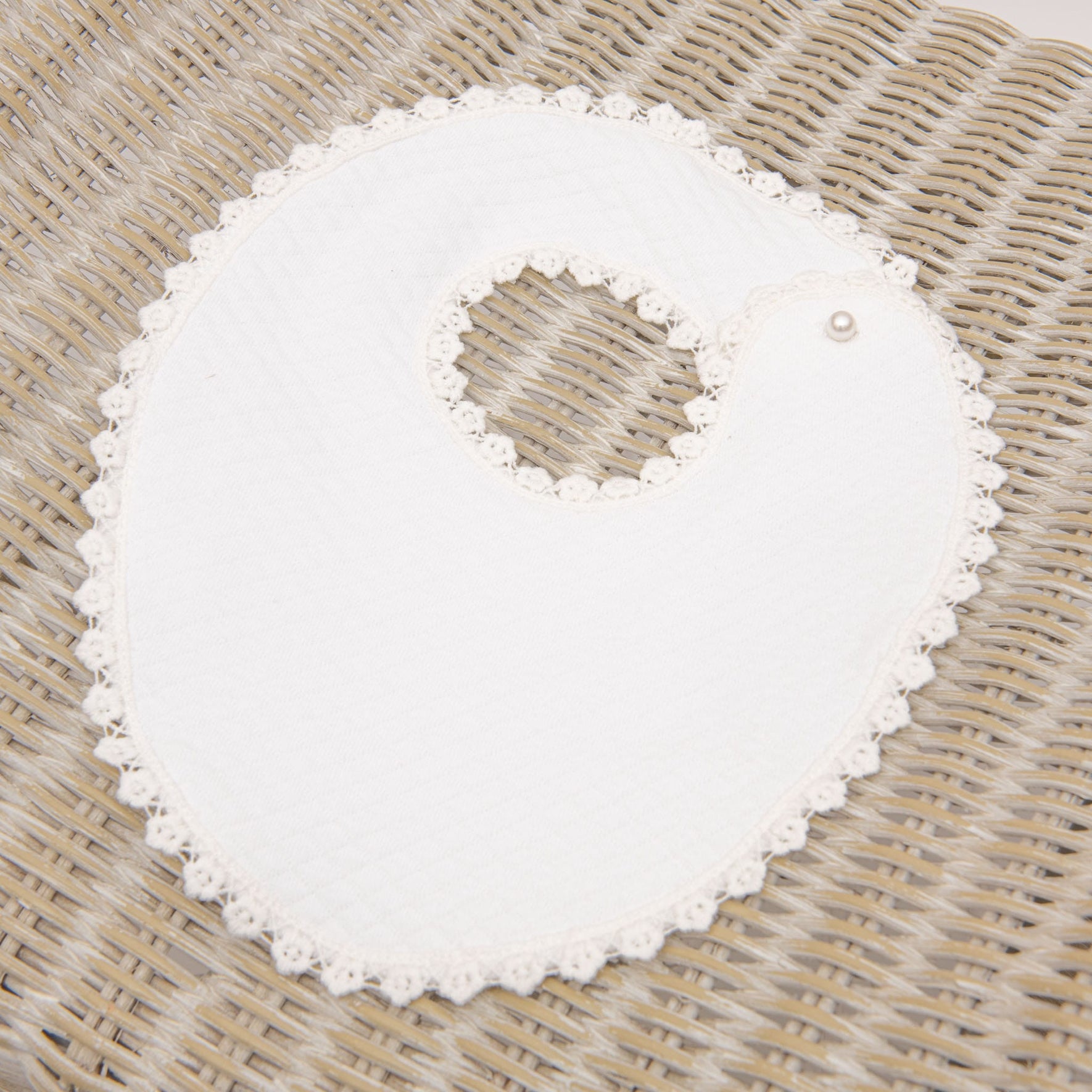 A white, round Eliza Bib with a floral lace border is lying on a woven, wicker surface. The bib has a circular neck opening and is fastened with a pearl-like button at one side, making it perfect for completing a baptism outfit.