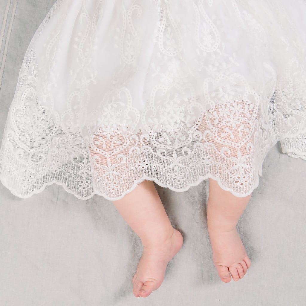 A baby wearing a handmade USA white lace Eliza Blessing Dress & Headband. The image captures the baby's lower body and bare feet. The intricate lace dress, made from luxurious Pima Cotton, extends slightly below the baby's knees. The baby is lying on a pale gray surface.