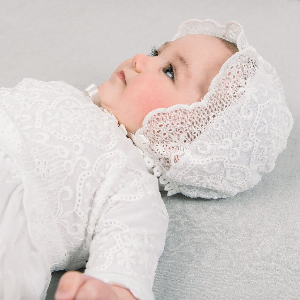 A baby girl wearing the Eliza Christening Bonnet and matching baptism gown lies on a soft gray background and looks up away from the camera in a thoughtful pose. 