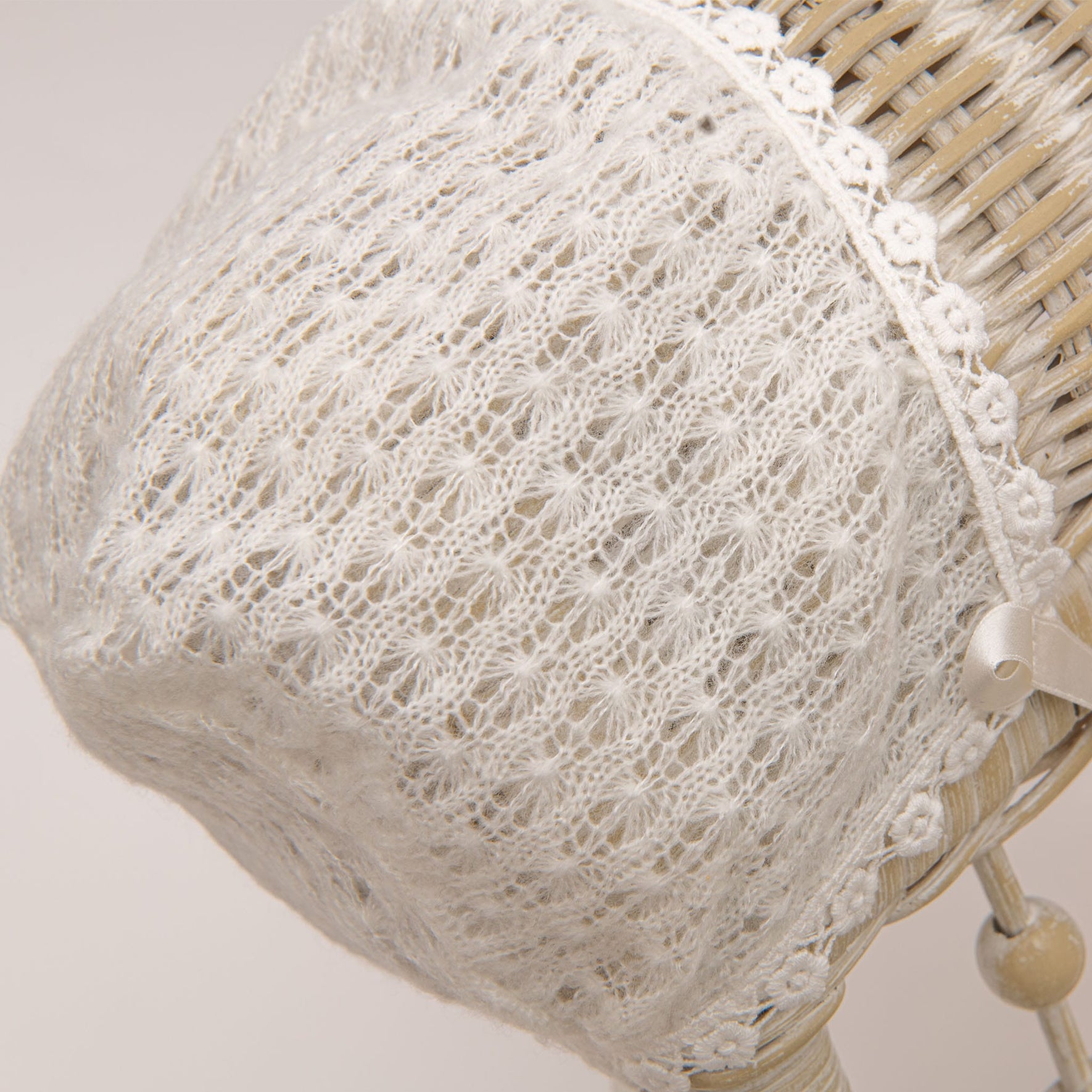 A close-up of a white lacy handmade Eliza Christening Knit Bonnet with intricate crochet patterns, placed on a light-colored wicker surface. The bonnet features a delicate floral trim and elegant silk ribbon ties on the side.