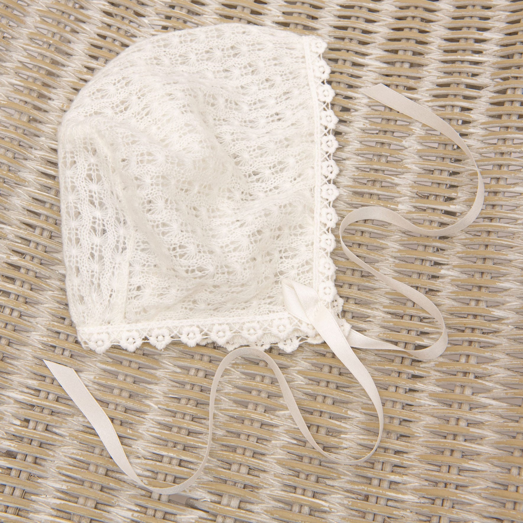 A delicate, white Eliza Christening Knit Bonnet with intricate patterns and a scalloped edge sits on a woven rattan surface. This handmade bonnet features two long silk ribbon ties for securing under the chin.
