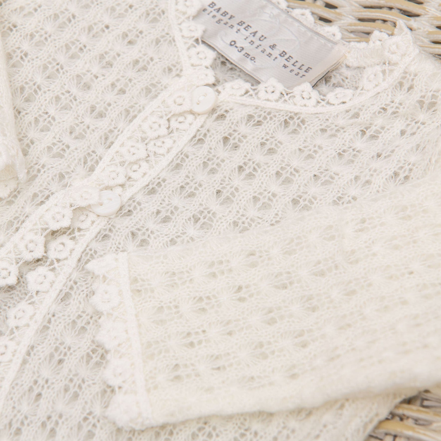 A close-up of a delicate, cream-colored lace cardigan featuring intricate patterns and small floral buttons. This Eliza Christening Knit Sweater by "Baby Beau & Belle" is partially visible near the neckline, indicating it is a size 3-6 months—a perfect Christening Day accessory.