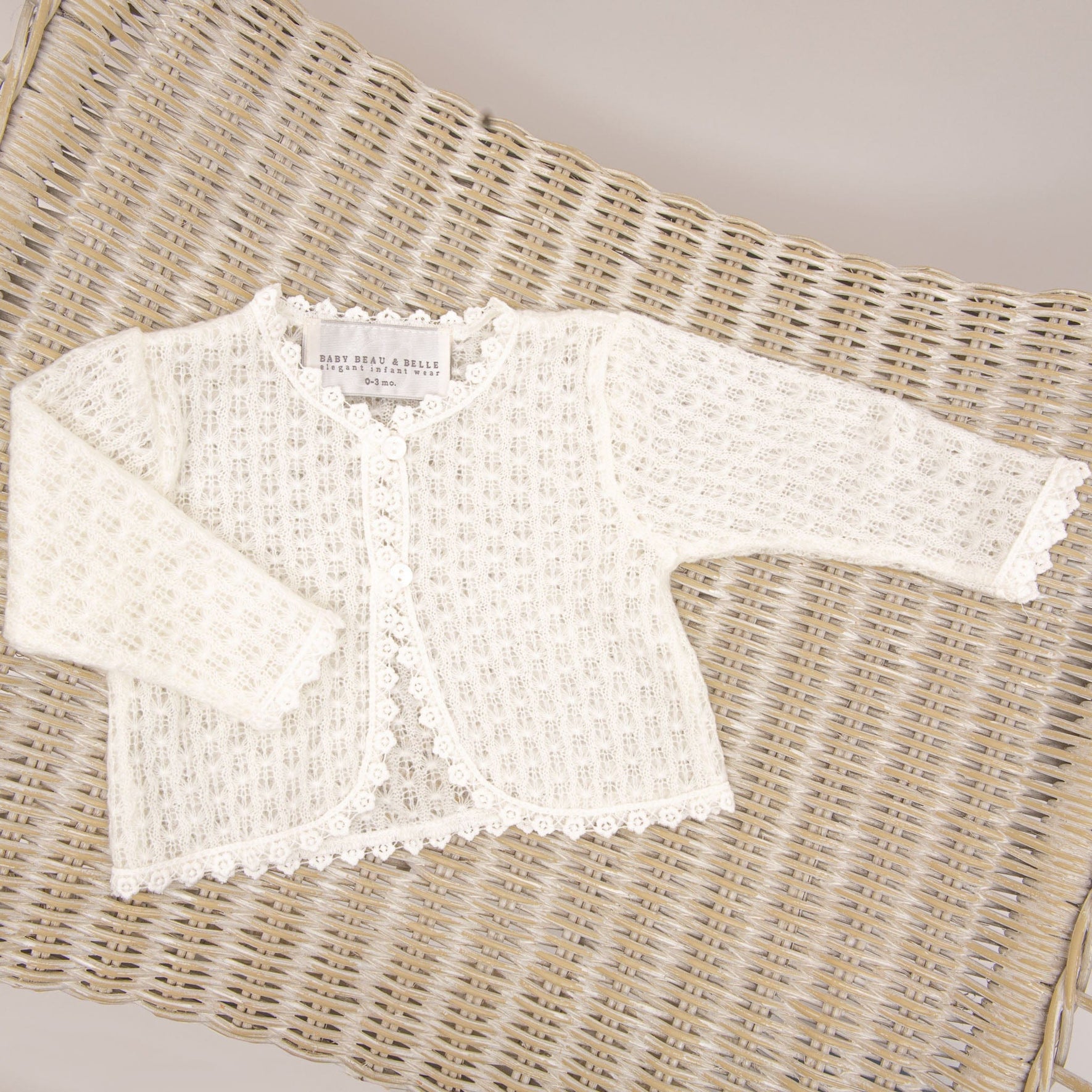 The image shows a long-sleeved, white lace Eliza Christening Knit Sweater with a scalloped edge, perfect as a Christening Day accessory. Displayed on a wicker surface, the label inside the collar reads "Baby Dior Paris" and includes additional text about size and care.