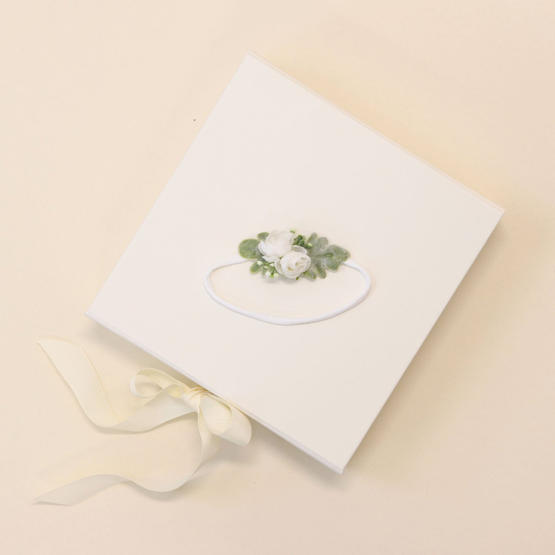 The Eliza Flower Headband is presented in a square white box adorned with a decorative floral design on the lid, showcasing two ivory flowers and green leaves. A cream ribbon elegantly ties the side of the box, which rests gracefully on a light beige surface, evoking the delicate craftsmanship of the handmade headband inside.