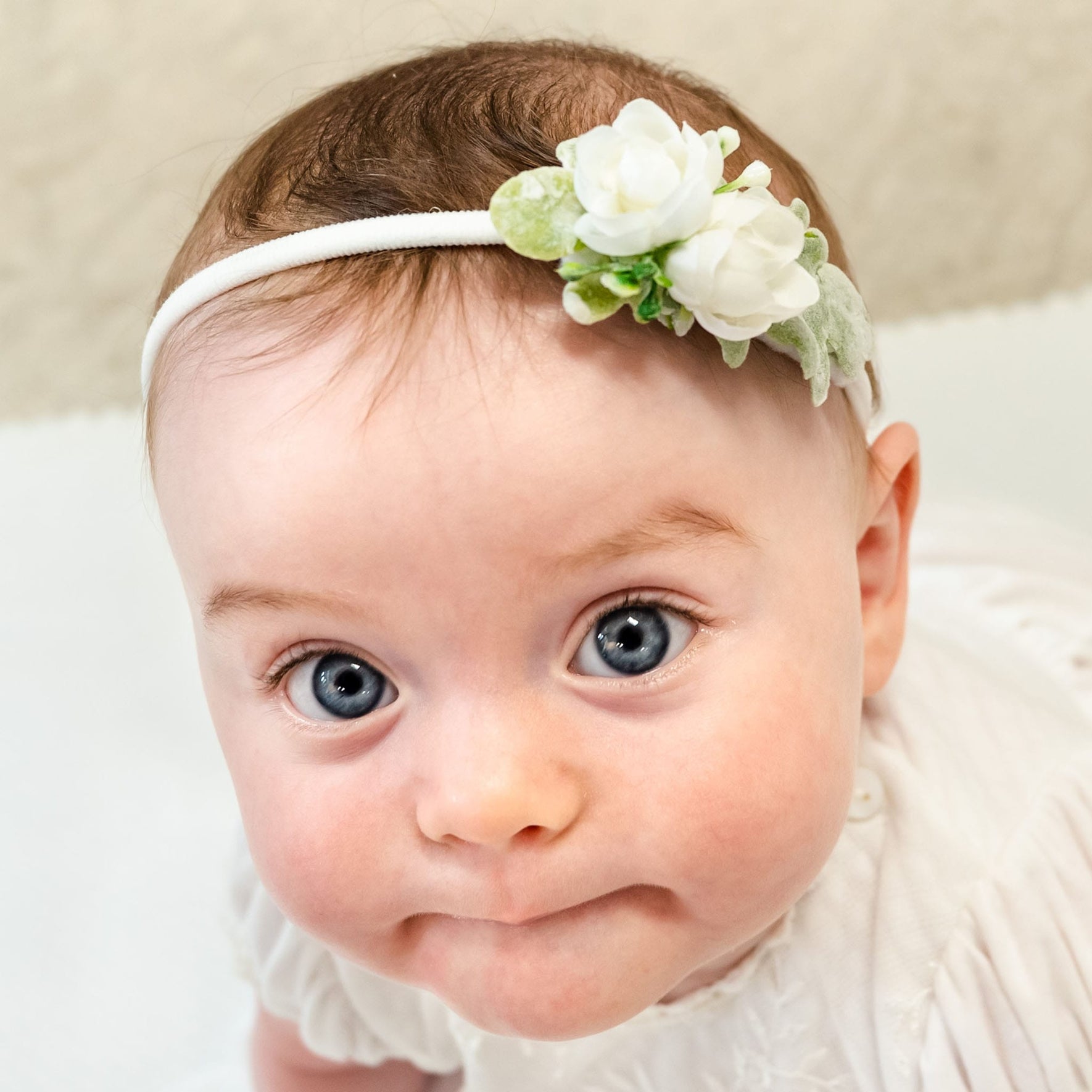 A baby with blue eyes wears the Eliza Flower Headband, which showcases ivory flowers on a soft nylon band. The baby looks upward with a neutral expression against a gentle beige backdrop.