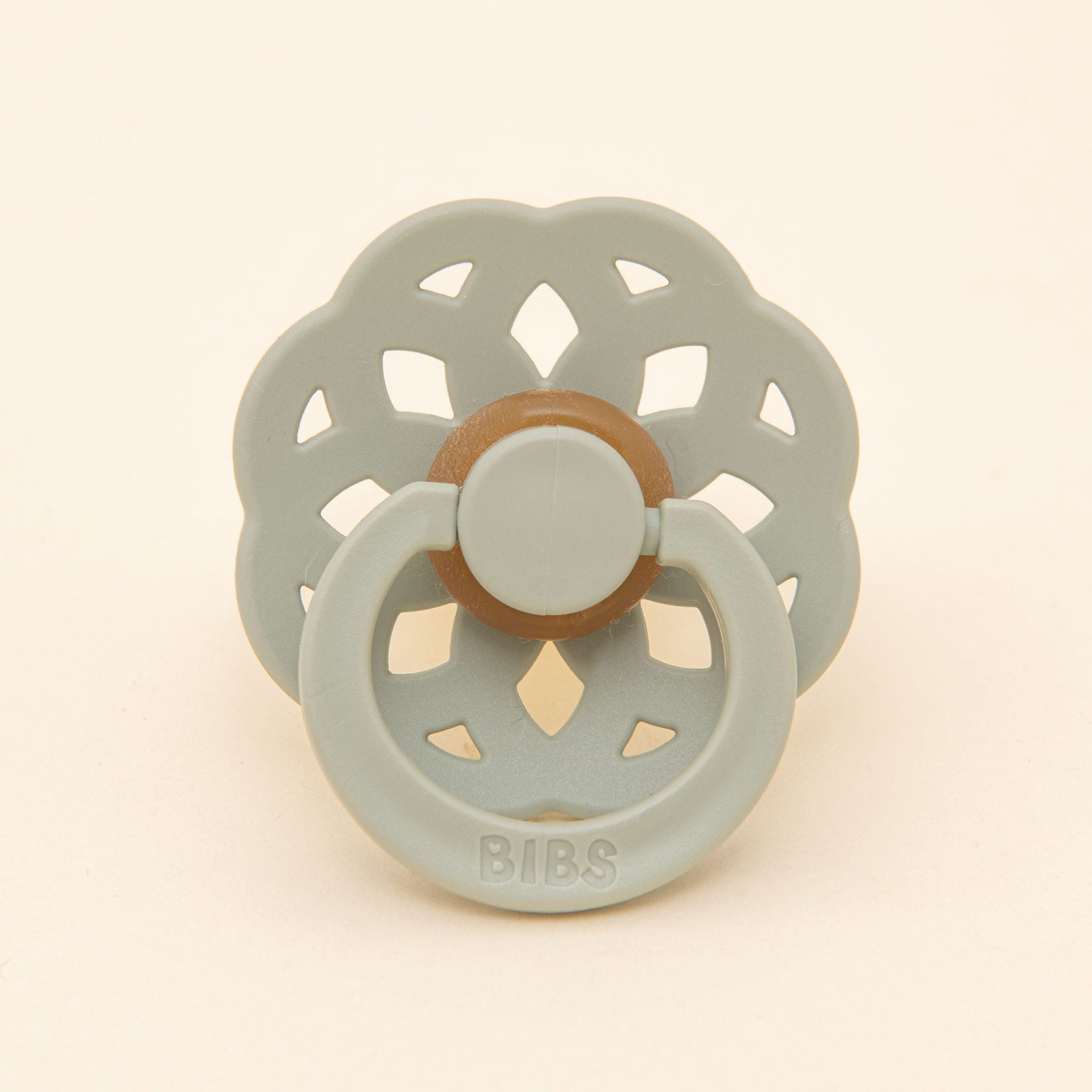 The Eliza Pacifier from the Sage & White set showcases a timeless and elegant design, featuring a floral-shaped shield and a round brown nipple crafted from natural rubber. Against a plain beige backdrop, it is complemented by the word "BIBS" embossed on the handle, making it perfect for helping little ones self-soothe.