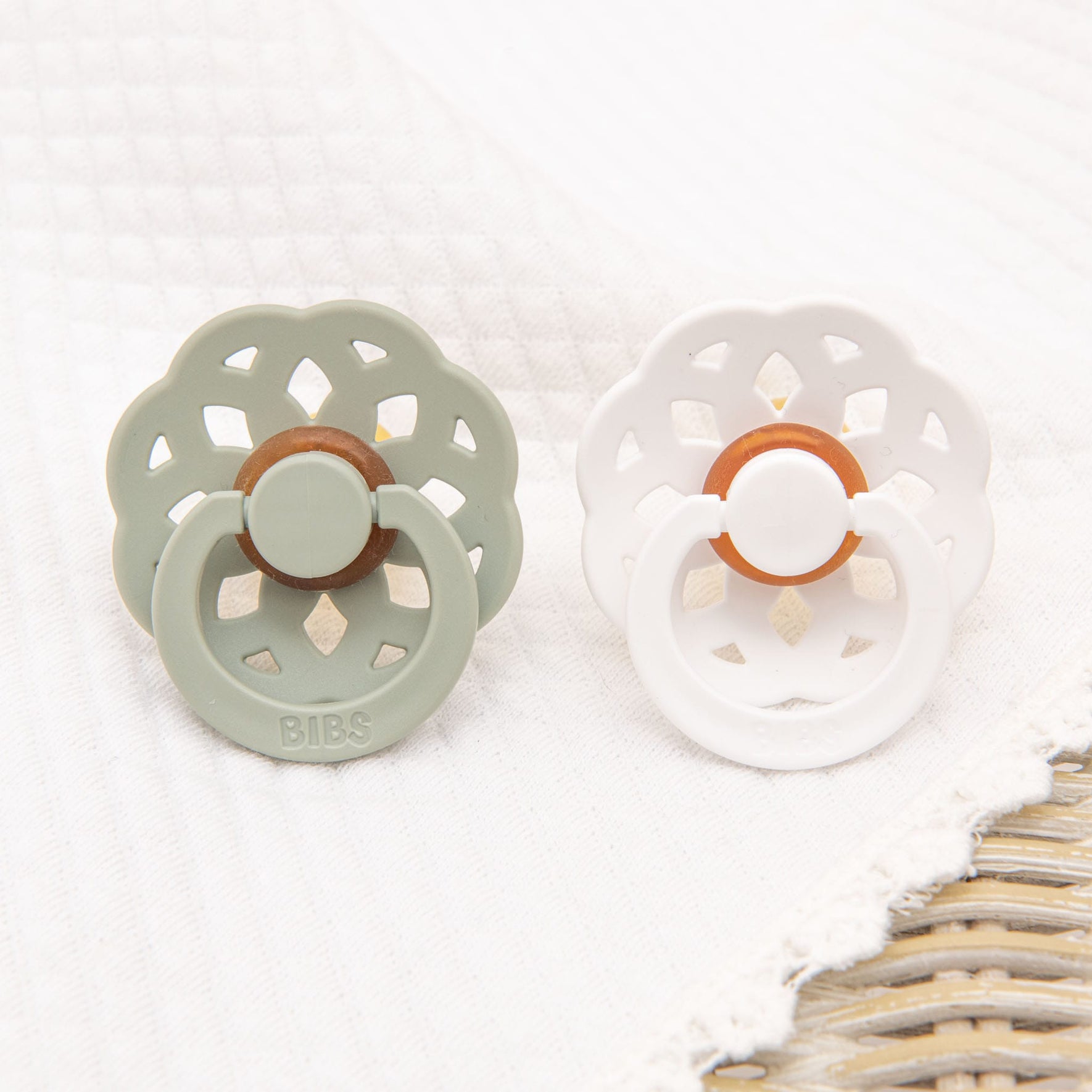 The Eliza Pacifier Set in Sage & White is displayed on a white textured surface, showcasing two pacifiers made from natural rubber. The sage pacifier features a decorative flower-like design, while the white one shares the same classic and elegant aesthetic. Both pacifiers include a central brown ring, perfect for aiding little ones in self-soothing.
