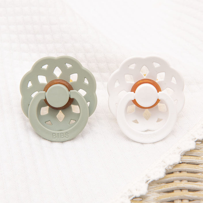 The Eliza Pacifier Set in Sage & White is displayed on a white textured surface, showcasing two pacifiers made from natural rubber. The sage pacifier features a decorative flower-like design, while the white one shares the same classic and elegant aesthetic. Both pacifiers include a central brown ring, perfect for aiding little ones in self-soothing.