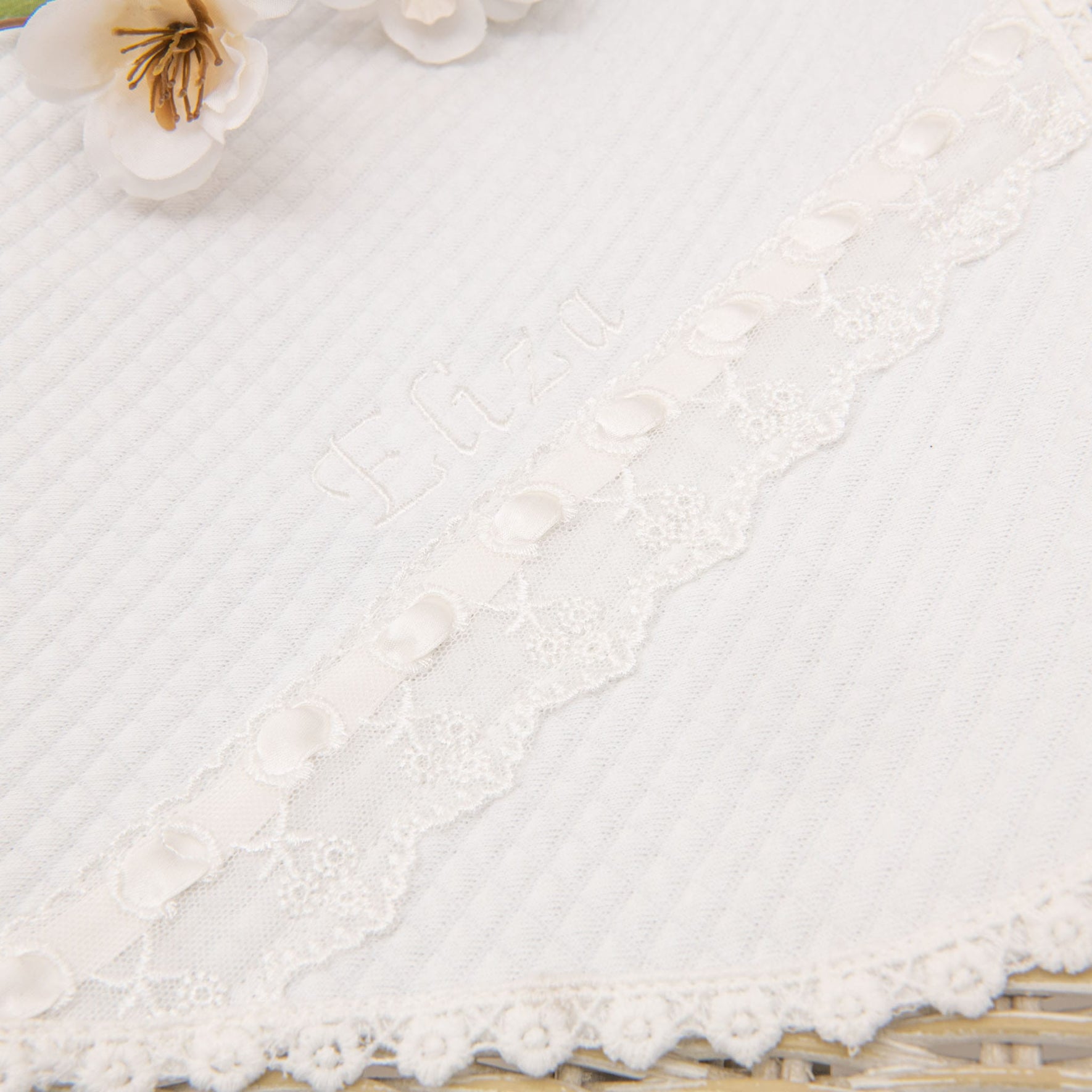 Close-up of a white textured fabric with embroidered flowers and floral lace trim. The product name "Eliza Personalized Blanket" is embroidered on the fabric, and there are decorative buttons along the lace edge. This handmade blanket appears to be a part of a baby blanket or similar delicate item.