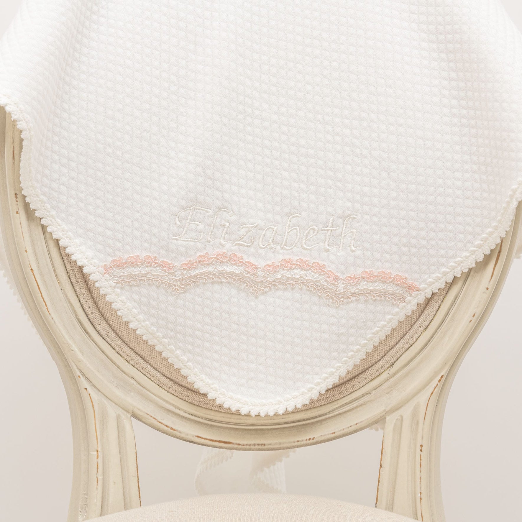 A white textured fabric, ideal for a christening outfit, with the name "Elizabeth" from the Elizabeth Accessory Bundle - Save 15%, is elegantly draped over the back of a vintage white wooden chair. The fabric is adorned with a delicate pink lace trim along the edge.