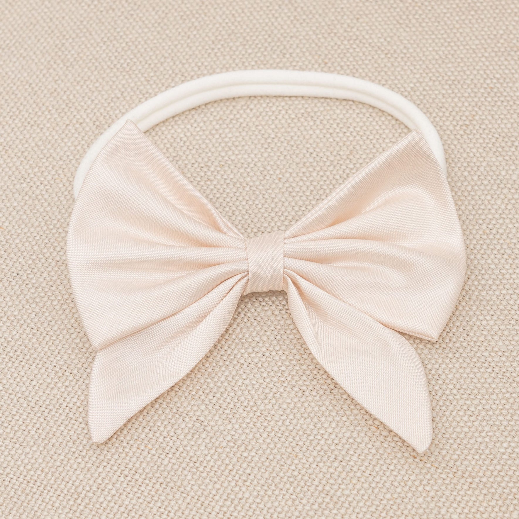 A pale pink fabric bow from the Elizabeth Accessory Bundle - Save 15%, perfect for a baptism dress, is attached to an elastic headband. The bow is neatly tied with two tails, and the headband lies on a textured beige surface.