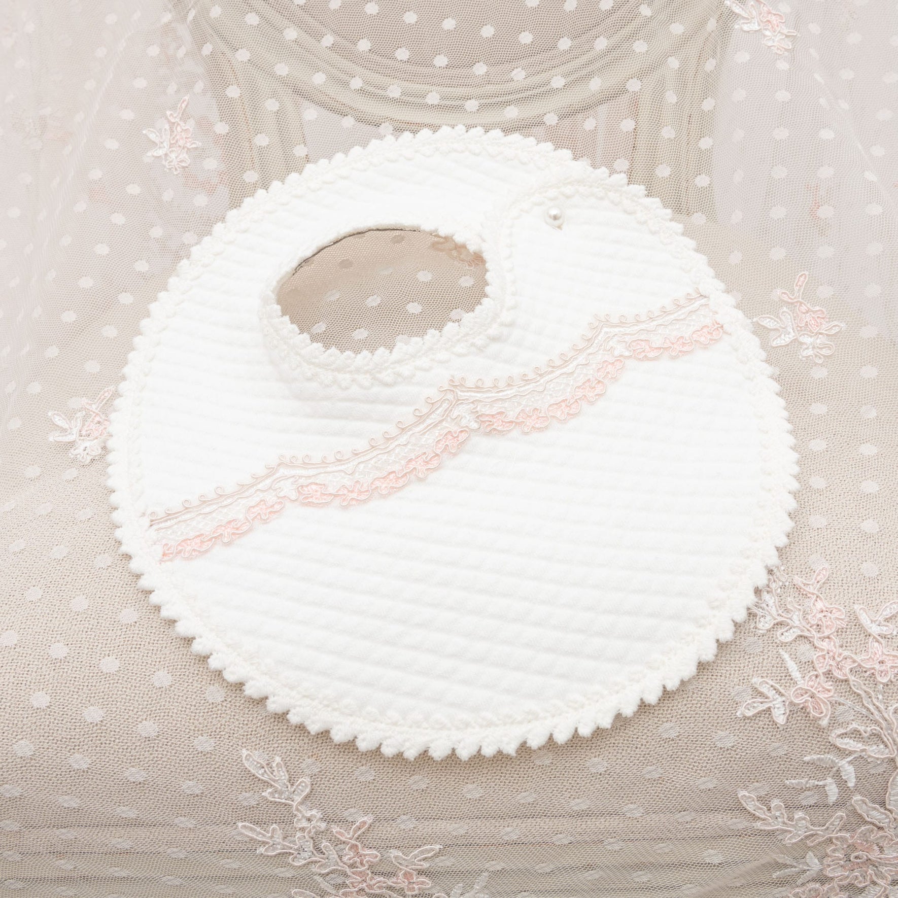 The Elizabeth Accessory Bundle, offering a 15% discount, features a textured white, round baby bib with a scalloped edge and pink lace detailing. It complements the sheer fabric adorned with white polka dots and pink floral embroidery, making it an ideal addition to a Christening outfit.