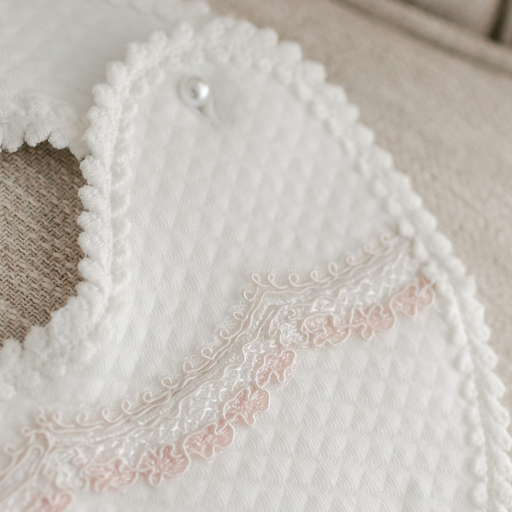 A close-up image of the Elizabeth Bib, featuring scalloped edges and delicate light pink embroidery details. The fabric showcases a textured diamond pattern, with a button snap at the top for fastening. Perfect for accentuating a special occasion dress, the bib is displayed against a soft, beige surface.