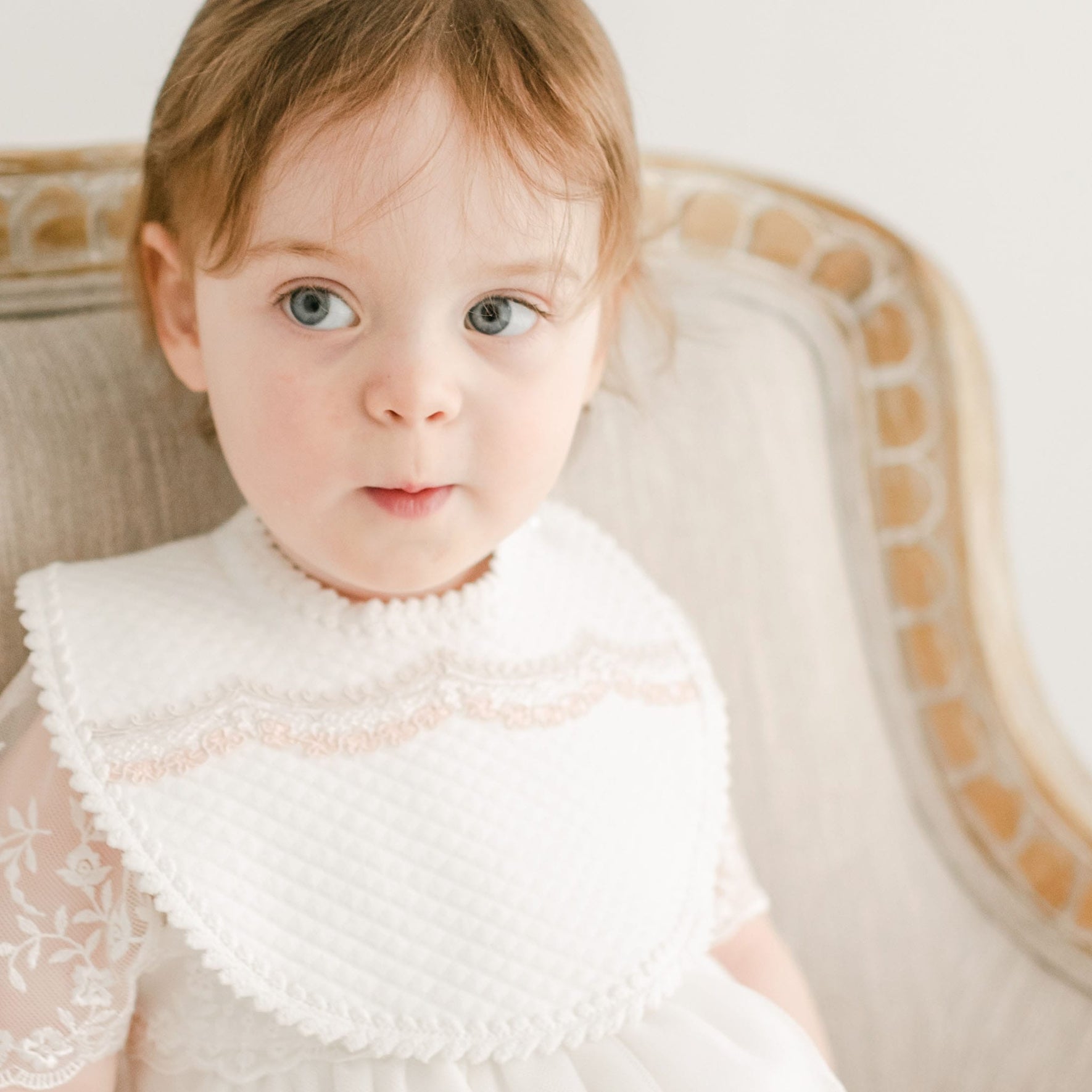 A toddler with light brown hair sits on a beige upholstered chair. Wearing a special occasion dress and the Elizabeth Bib, which features handmade lace detailing, the child looks slightly to the side with an expression of surprise or curiosity, adding a charming vintage touch to the scene.