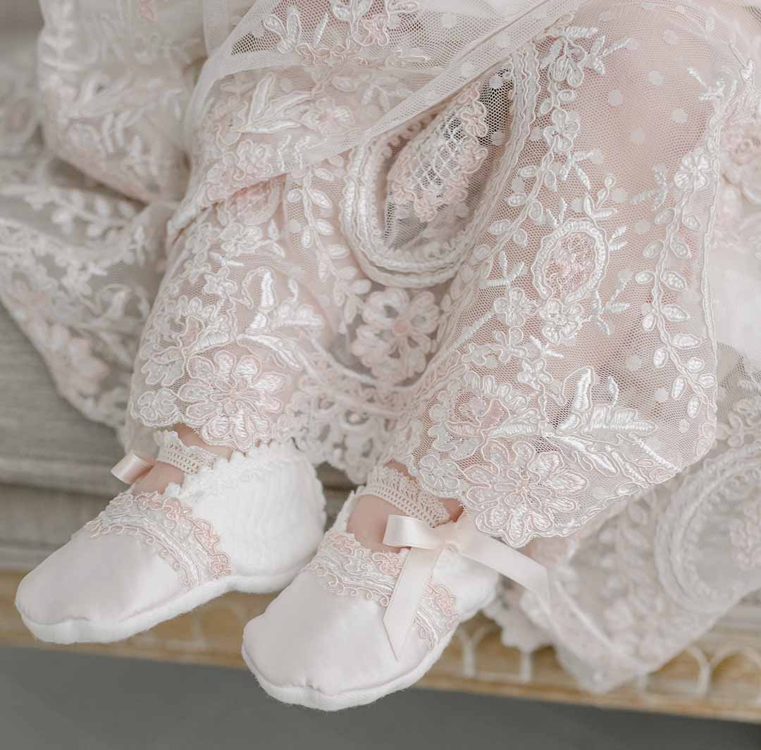 A baby is wearing an intricate, lace-patterned christening gown adorned with floral designs and the delicate Elizabeth Booties, which are accented by pink silk ribbons. The image highlights the exquisite details and textures of the fabric.