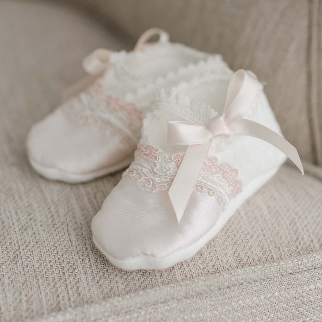 A pair of Elizabeth Booties, soft and light pink with lace detailing and small pink silk ribbons, rests on a beige textured fabric surface.