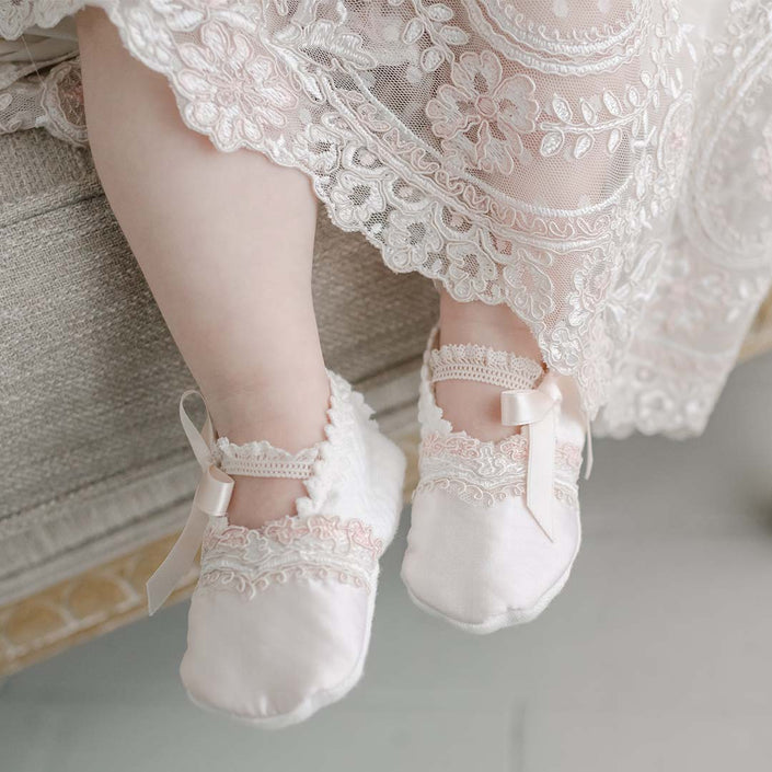A baby wearing Elizabeth Booties, adorned with pink silk ribbons and lace trim, is sitting on a beige surface. The baby is dressed in a lacy, patterned fabric reminiscent of a christening gown. The scene is softly lit, highlighting the delicate textures of the clothing and booties.