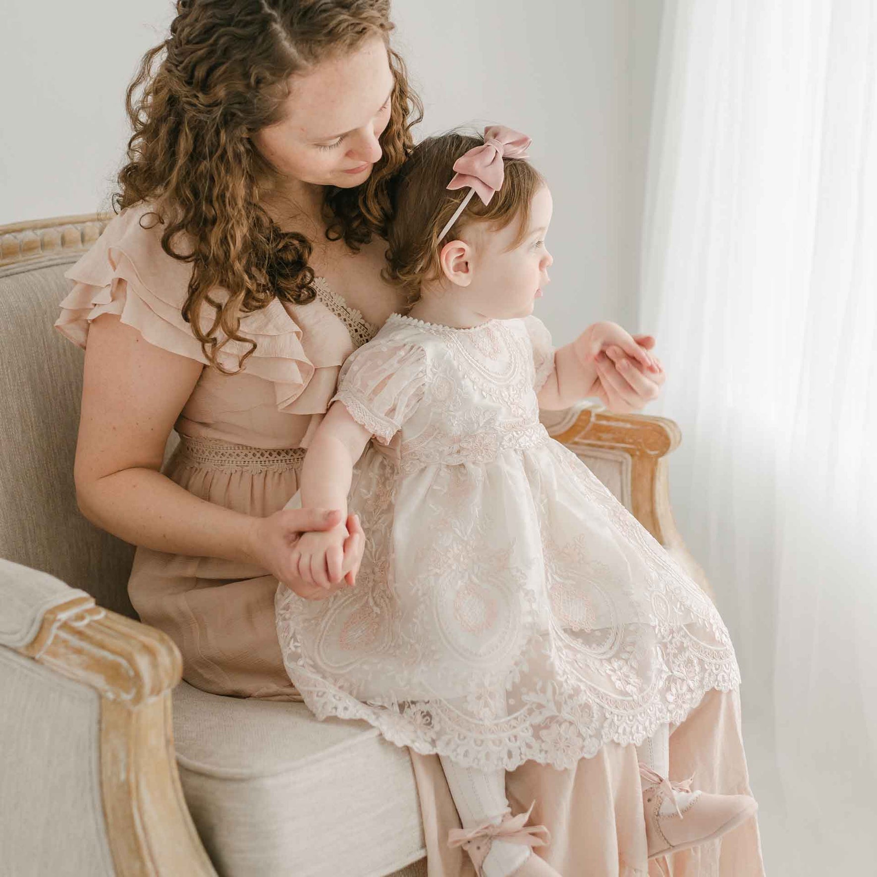 A woman with curly hair, wearing a light pink dress, sits on a light-colored armchair while holding a young child. The child, dressed in the Elizabeth Christening Dress and a pink bow headband, looks out towards the window. Both appear peaceful and focused.