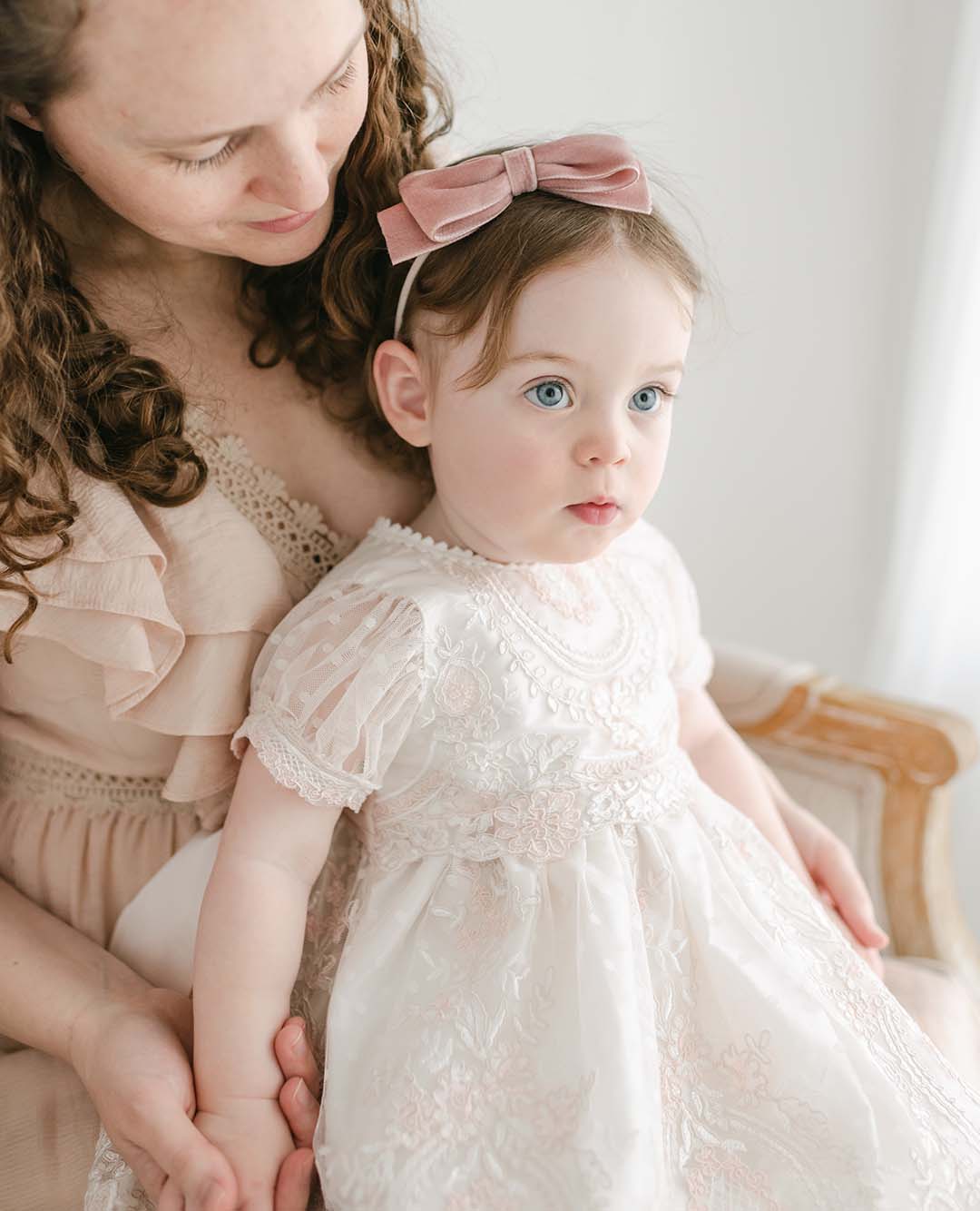 A woman with curly hair smiles as she holds a young girl on her lap. The girl is wearing the Elizabeth Christening Dress, a pale pink lace baptism dress paired with a bow headband. They are seated on a wooden chair with a light-colored cushion. The background is bright and softly lit.