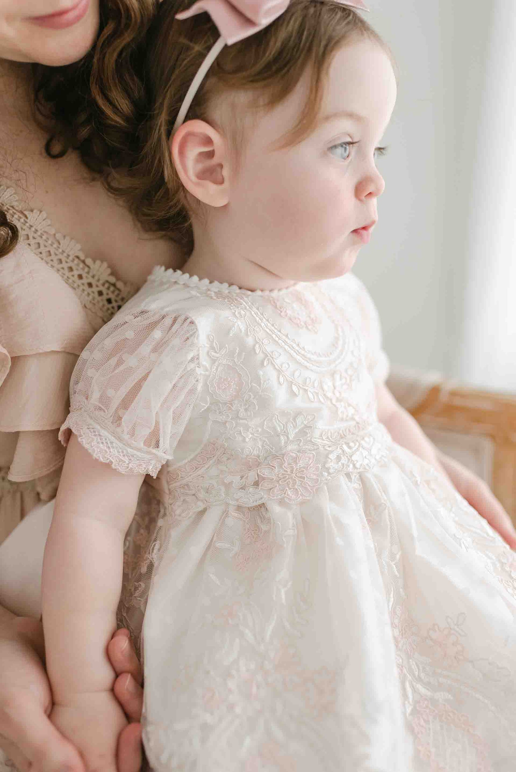 A toddler with light brown hair, wearing the Elizabeth Christening Dress adorned with delicate floral patterns, sits in the arms of an adult. The child looks to the side with a thoughtful expression, and both figures are partially visible in the softly lit scene.