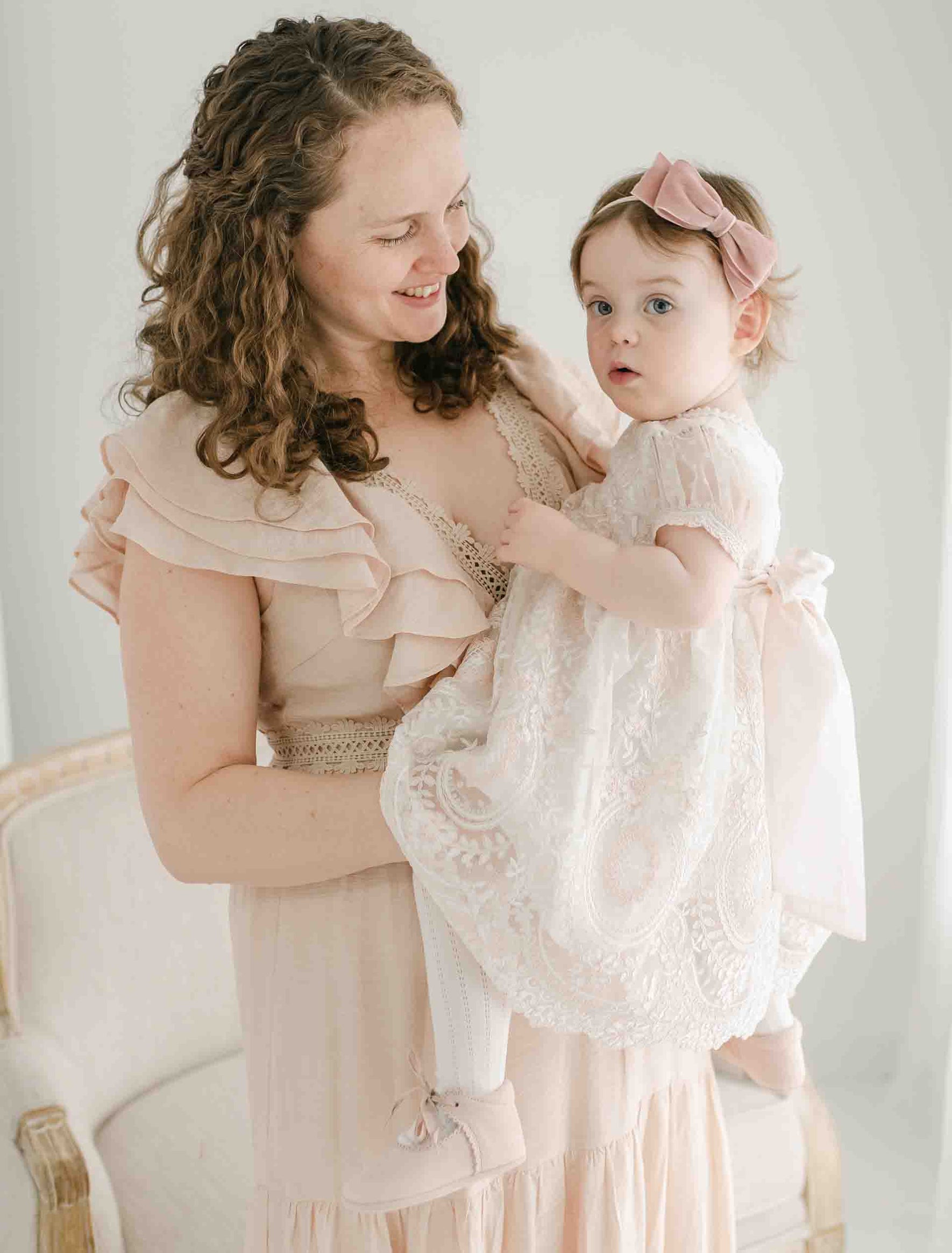 An adult with curly hair in a handmade light-colored dress is holding a young child dressed in the Elizabeth Christening Dress with a pink bow on her head. They are standing in front of a pale background with a chair visible to the left. The adult is smiling while looking at the child.